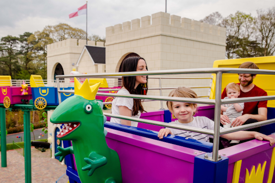 Photo of a family enjoying a theme park ride with a purple dinosaur character and a castle in the background.