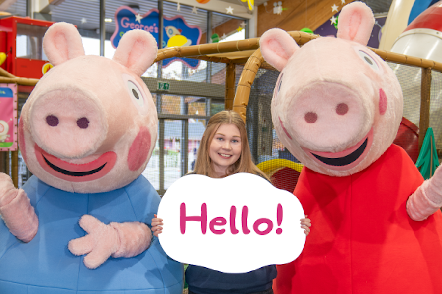 Photo of a young girl holding a 'Hello!' sign standing between two animated pig characters in an indoor play area.